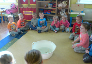 Przedszkolaki siedzą w kole, w środku stoi na dywanie miska z wodą, w której leżą drobne monety.
