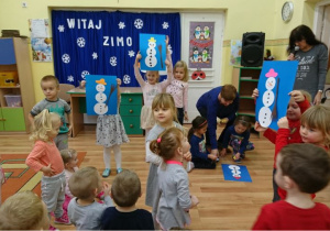 3 pary przedszkolaków przyklejają na niebieskie kartony sylwety bałwanków z gotowych elementów.