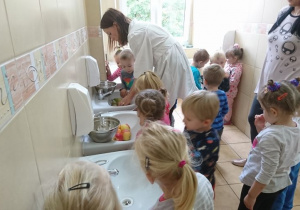 Dzieci z pomocą nauczycielki myją owoce w łazience.