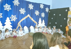 Dzieci w przebraniu aniołków siedzą w kole na tle bożonarodzeniowej dekoracji.
