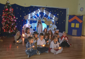 Grupa przedszkolaków siedzi na podłodze z zapalonymi świeczkami, jedna z dziewczynek śpiewa kolędę.