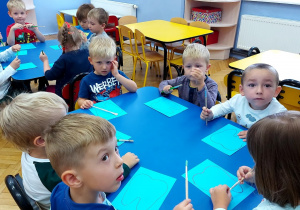 Dzieci siedzą przy stołach i trzymają w rączkach pędzle do malowania farbami.