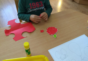 Chłopczyk siedzi przy stoliku i przykleja skrawki czerwonego papieru na szablonie jabłka.