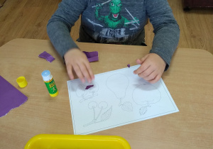 Przedszkolak wykleja fioletowymi skrawkami papieru szablon śliwki.