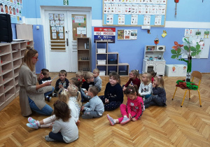 Grupa dzieci siedzi na podłodze i słucha opowiadania nauczycielki.