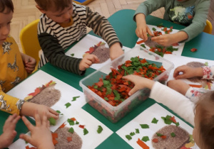 Dzieci ozdabiają przy stoliku sylwety jeżyków za pomocą ścinków materiału.
