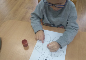 Chłopczyk maluje kontur misia brązową farbą plakatową za pomocą patyczka.