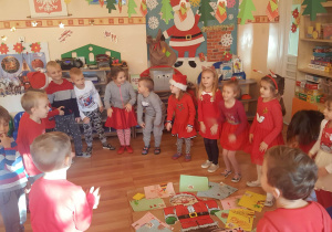 Dzieci stoją w kole wokół postaci św. Mikołaja, kładą obok rysunki prezentów, o których marzą.