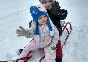 2 dzieci siedzi na sankach i macha rączką do pozostałych.