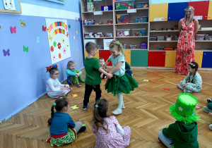 Chłopczyk tańczy w parze z dziewczynką, pozostałe dzieci przyglądają się im. Przed nimi leżą motyle w kolorach: żółtym, pomarańczowym, zielonym, niebieskim, różowym