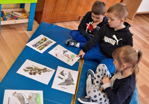 Dwóch chłopców i jedna dziewczynka siedzą na materacach. oglądają obrazki z ptakami i kwiatami zwiastującymi nadejście wiosny.