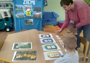 Przedszkolaki z pomocą nauczycielki segregują obrazki odpadów do właściwych pojemnikow.