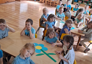 Chłopcy i dziewczynki siedzą przy stolikach. Układają kwiat niezapominajki w małych zespołach, z niebieskich, żółtych i zielonych elementów z kartonu.