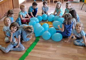 Grupa 4 - 5 latków ułożyła na podłodze duży kwiat niezapominajki, składający się z błękitnych balonów, oraz z klocków w kolorze żółtym i zielonym.