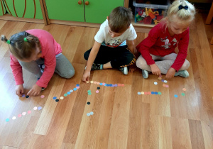 Dzieci siedzą na podłodze. Układają z kolorowych kółek obrazki według swojej wyobraźni.