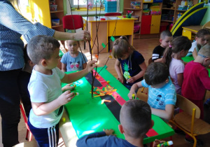 Dzieci przy stole ozdabiają kolorowym papierem drzewka na makiecie.