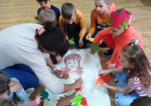 Przedszkolaki razem z panią Emilką ozdabiają kolorowymi listkami postać Pani Jesieni.