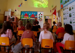 Dzieci siedzą na krzesełkach. Na ścianie wyświetlony jest film pt. „Piotruś Królik”