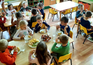 Dzieci siedzą przy stolikach. Na stołach postawione są talerze z bananami, winogronem, pączkami, oraz babeczkami. W kubeczkach dzieci mają wodę.