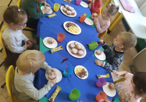 Dzieci siedzą przy wspólnym stole, jedzą słodkości przygotowane przez mamy przedszkolaków.