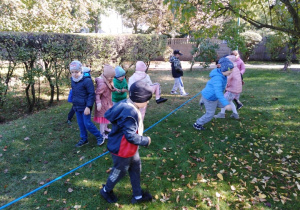 Przedszkolaki przeskakują przez rozciągniętą na wysokości kolan niebieską linę.