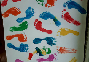 Zdjęcie kartonu, na którym odbite są pomalowane farbą stopy dzieci i nauczycielki w kolorach: niebieskim, żółtym, czerwonym, zielonym, pomarańczowym i różowym.