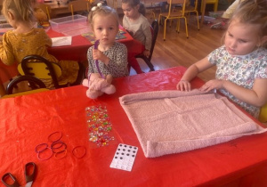 Dwie dziewczynki przy stoliku - jedna trzyma wykonaną maskotkę, druga składa ręcznik według wzoru.