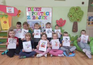 Grupa przedszkolaków siedzi na tle dekoracji z dużym kolorowym napisem: "Dzień postaci z bajek" z rysunkami w rączkach.