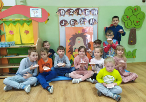 Grupa dzieci siedzi na tle dekoracji z napisem "Dzień Jeża" i postacią dużego jeża. Przedszkolaki trzymają w rączkach własnoręcznie wykonane jeżyki z masy solnej i wykałaczek.