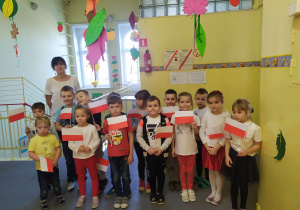 Grupa starszaków razem z panią Emilką stoi na przedszkolnym korytarzu. Dzieci są w odświętnych ubraniach i trzymają biało - czerwone chorągiewki.