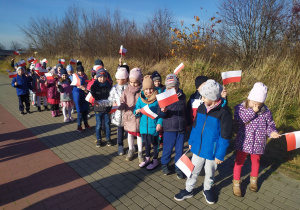 Dzieci z grup: "Słoneczka" i "Misie" spacerują w okolicy przedszkola z biało - czerwonymi chorągiewkami w rączkach.
