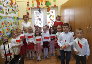 Ubrane na galowo 5-latki z grupy ""Misie" stoją z flagami w ręku na holu przedszkola.