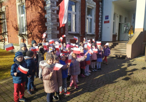 Przedszkolaki z grupy "Słoneczka" i "Misie" stoją przed wejściem do przedszkola z flagami w rączkach.