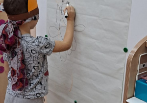 Chłopiec z zasłoniętymi oczami stoi przed tablicą i rysuje misia.