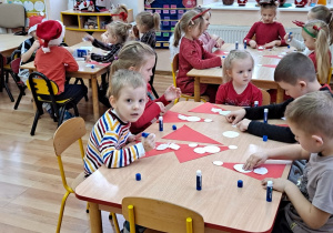 Cała grupa siedzi przy stolikach i tworzy Mikołajka z czerwonego trójkąta i białych kółek.