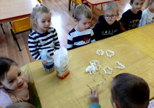 Dzieci siedzą przy stołach. Na stole stoi mąka i cukier. Dzieci poznają produkty potrzebne do zrobienia pierników.