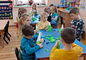Dzieci siedzą przy stolikach, owijają styropianowe stożki zieloną bibułą, oraz ozdabiają je cekinami i gwiazdkami z brokatowego papieru.