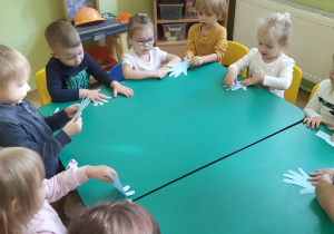 Przedszkolaki siedzące przy złączonych stolikach przymierzają wycięte szablony dłoni do swoich