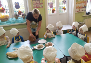 Dzieci siedzące przy wspólnym stole ubrane w fartuchy i czapki kucharskie, ciocia w fartuchu za pomocą noża do chleba kroi go na kromki.