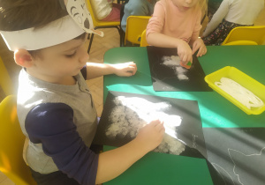 Przedszkolaki podczas wykonywania pracy plastycznej, na czarnym kartonie wypełniają kontur kundelka białą farbą nakładają ją gąbką.