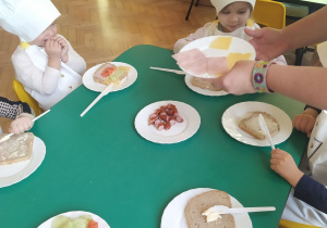 Dzieci wraz z panią Moniką układają wybrane składniki wędlinę, ser żółty na chleb