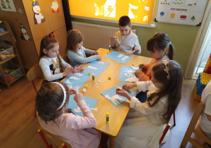 Grupa dzieci siedzi przy stoliku i wykleja zimowy obrazek z waty i białej bibuły.