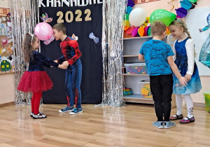 Dwie pary przedszkolaków poruszają się w rytm muzyki, utrzymując balony między czołami.