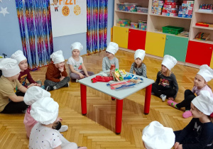 Dzieci siedzą wokół stolika, na którym znajdują się składniki do przygotowania pizzy