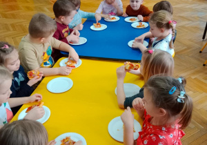 Dzieci przy stolikach jedzą upieczoną pizzę.
