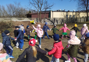 Dzieci na ogrodzie przedszkolnym maszerują w kole i śpiewają piosenkę pt. "Maszeruje wiosna".