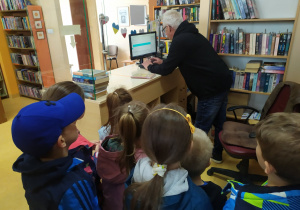 Pracownik biblioteki przy komputerze wyjaśnia dzieciom zasady wypożyczania książek.