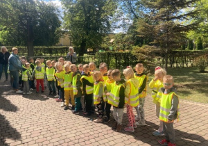 Dzieci z grupy Biedronek i Misiów ubrane w kamizelki odblaskowe ustawione przed budynkiem przedszkola czekające na wyjście na skrzyżowanie.