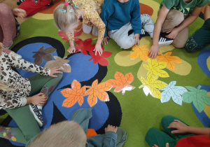 Dzieci tworzą jesienną kompozycję z liści rozłożonych na dywanie.