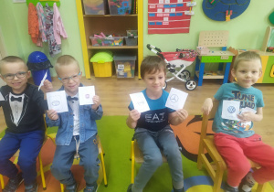 4 przedszkolaków siedzi w rzędzie na krzesłach i trzyma kartoniki z logo marek samochodów osobowych.